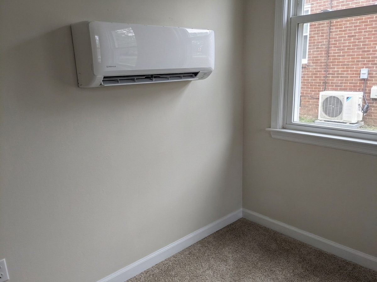 A white wall-mounted ductless mini-split air conditioning unit in a room corner with beige walls, carpet, and a window showing an outdoor condenser unit against a brick building.