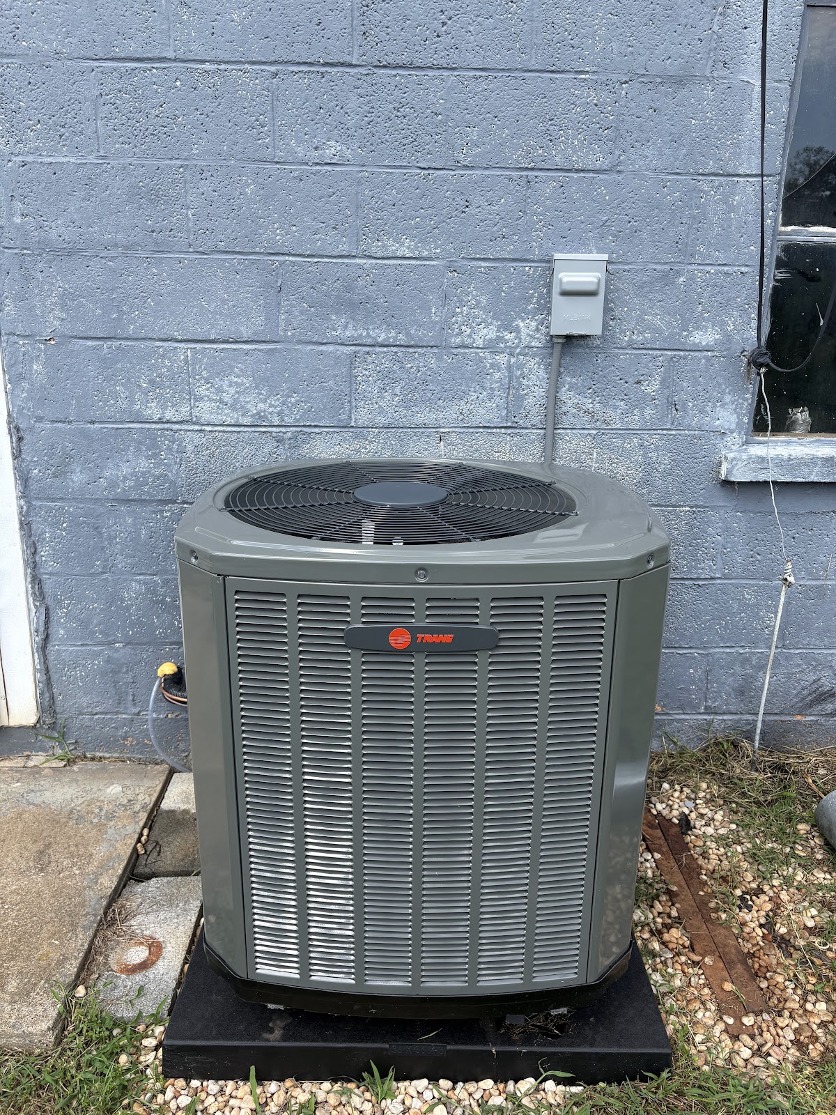 A Trane outdoor air conditioning condenser unit sitting on a black pad next to a gray cinder block building wall with an electrical disconnect box mounted above it.